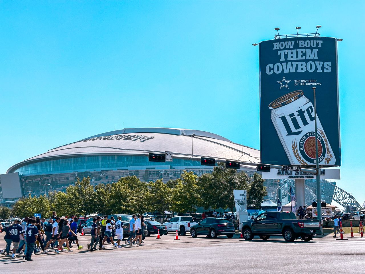 services-01 People walking near AT&T Stadium on a sunny day in Arlington, Texas, with a prominent billboard.