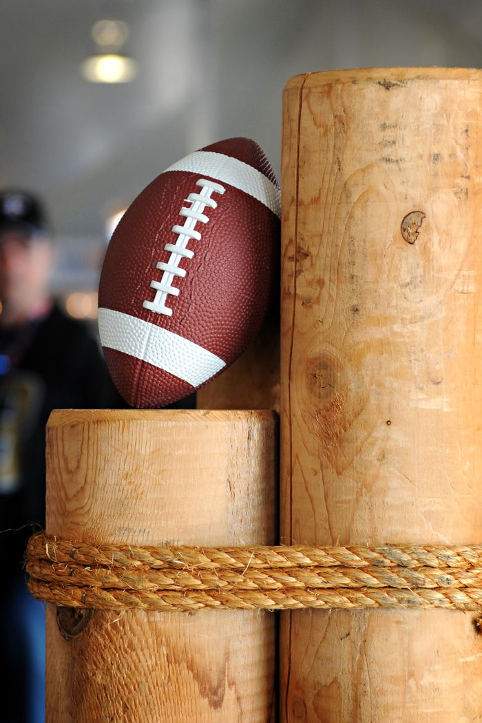 gallery-5 Close-up of a football resting on stacked wooden logs with a rustic rope indoors.